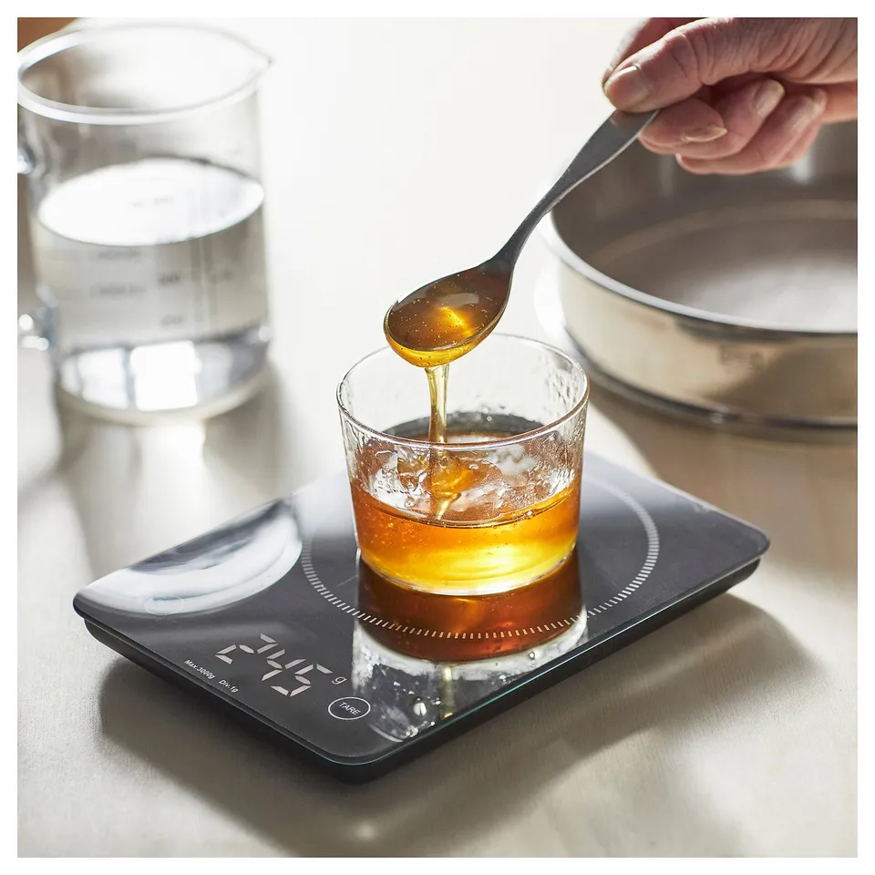 A person is pouring honey into a glass on a digital scale, GENOMSNITT, which is black.