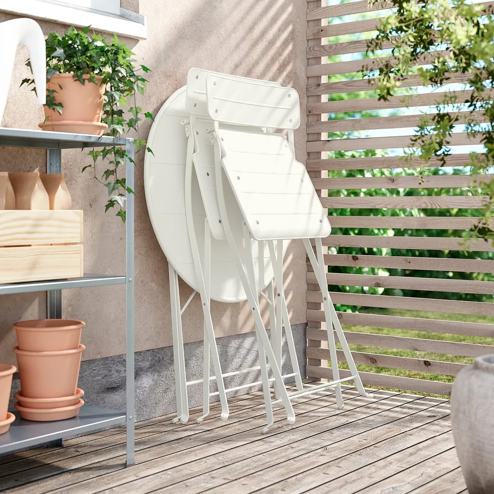 Three white SUNDSÖ folding chairs stacked on wooden deck, surrounded by potted plants and a wooden fence.
