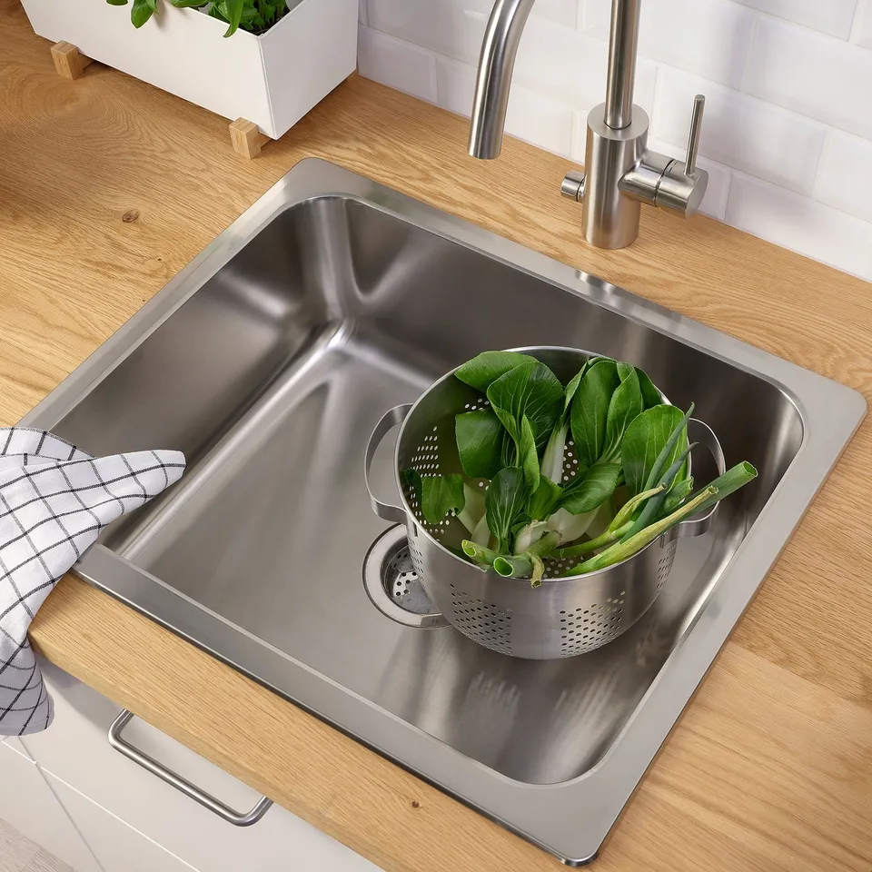 Stainless sink with strainer full of green veggies, wooden countertop, modern tap.