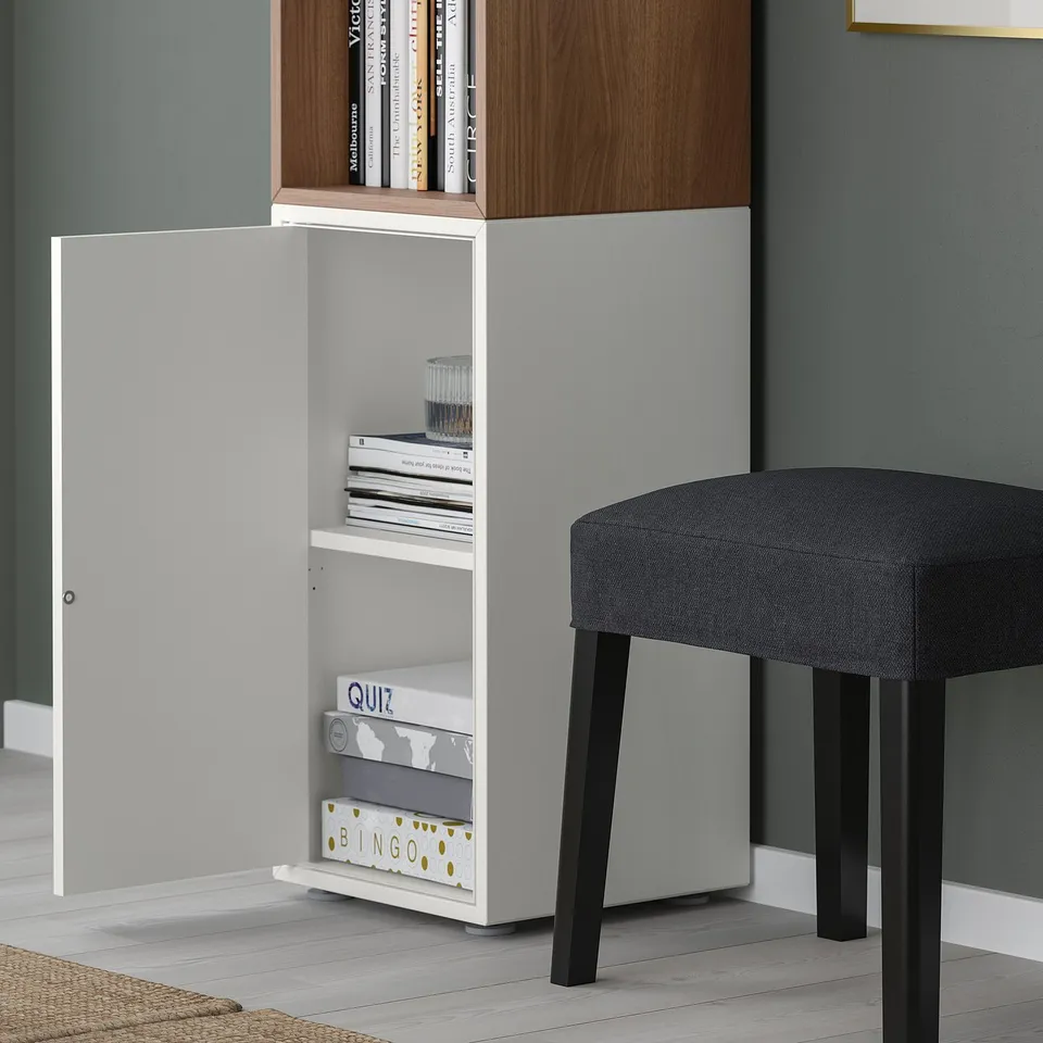 White EKET storage cabinet with open door revealing shelf holding game boxes and books. Black stool beside it.