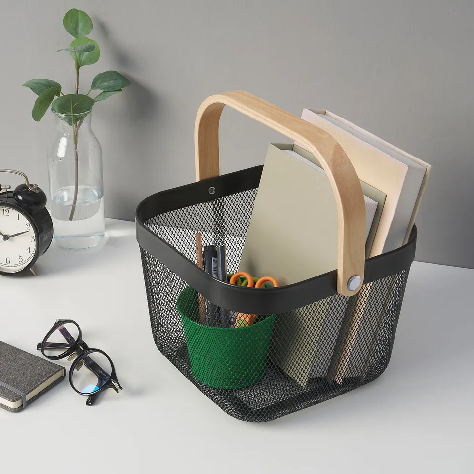 Black mesh storage basket with wooden handle, holding folders and office supplies, on a desk with a plant and clock.