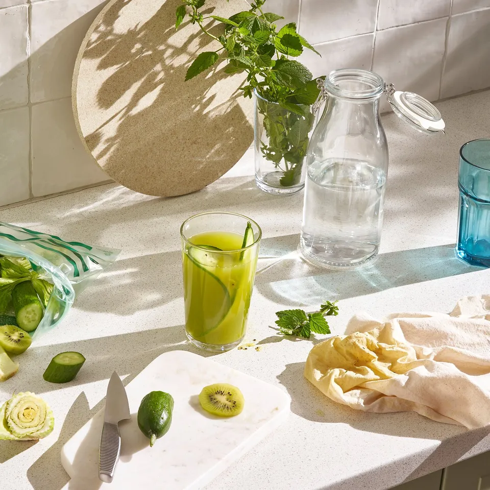 Countertop with KORKEN jars, one storing herbs, another with water, and a glass of green juice beside a cutting board with kiwi and limes.