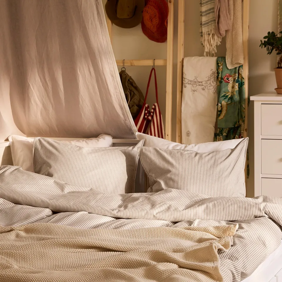 Bed with SOLFIBBLA striped beige & white duvet cover & pillows, light fabric, neutral decor, hanging items, small plant.