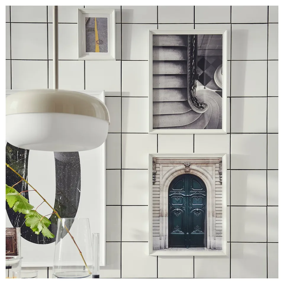 Three framed photos hang on a white tiled wall: a road line, a spiral staircase, and a blue door. A circular mirror with a plant sits beside them.