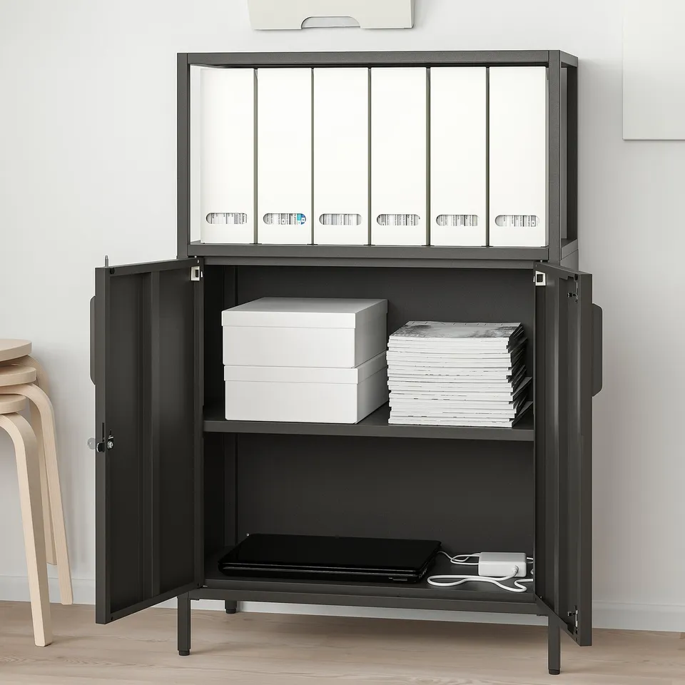 Black TROTTEN storage unit with adjustable shelves, lockable doors, and wall fastener. Holds folders, books, and a tray with chords.
