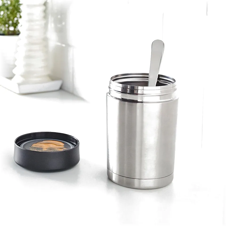 Silver thermos with a spoon, containing soup, beside its lid on a white countertop.