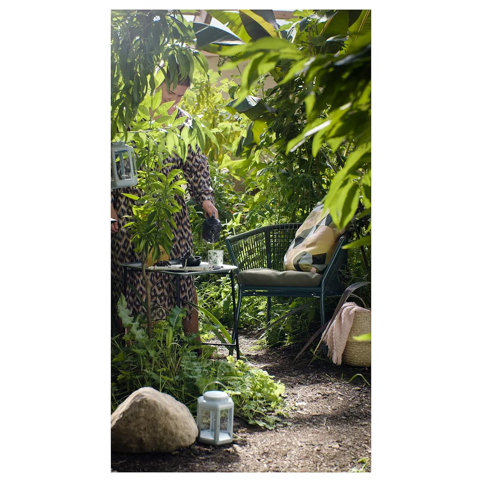 Person sitting in garden, surrounded by lush greenery, with a green SEGERÖN steel and rope chair.