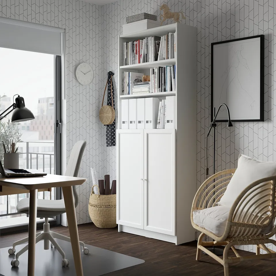 Modern home office with a white BILLY bookcase filled with books, next to a round wicker chair with a cushion, near an office desk with a lamp.