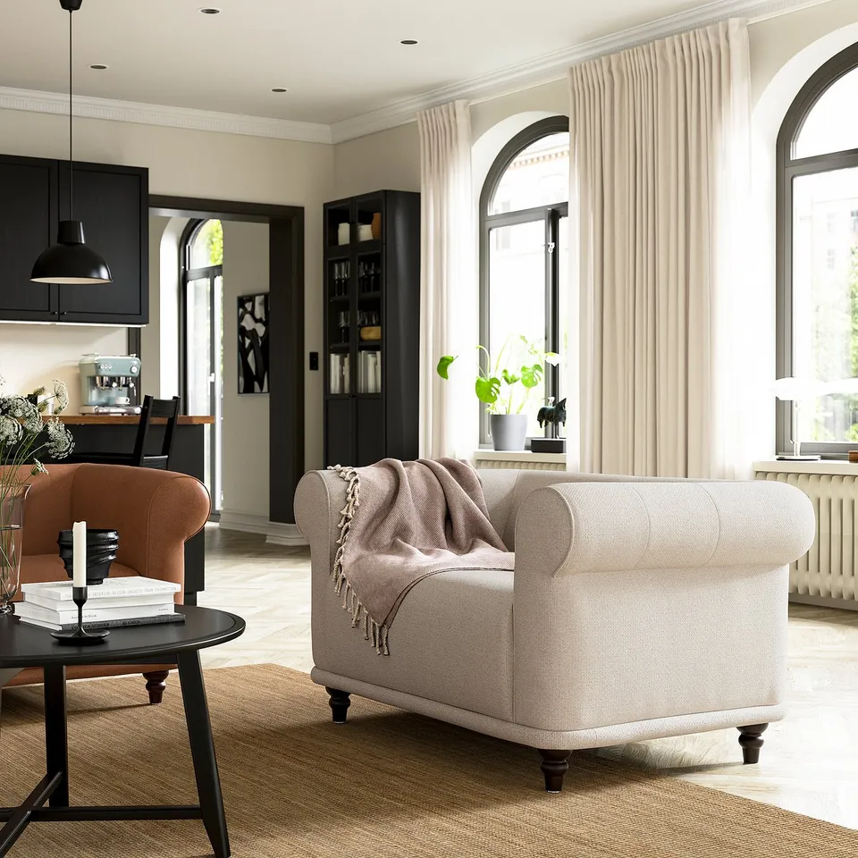 Cosy living room with a beige VISKAFORS armchair, wooden coffee table, and bookshelf. Soft lighting, curtains, and plants.