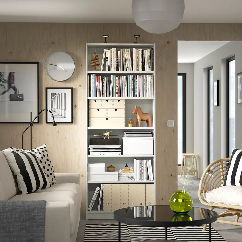 Cosy living room: beige sofa, striped pillows, bookcase, coffee table, green vase, rug, clock, lamp, wicker chair.