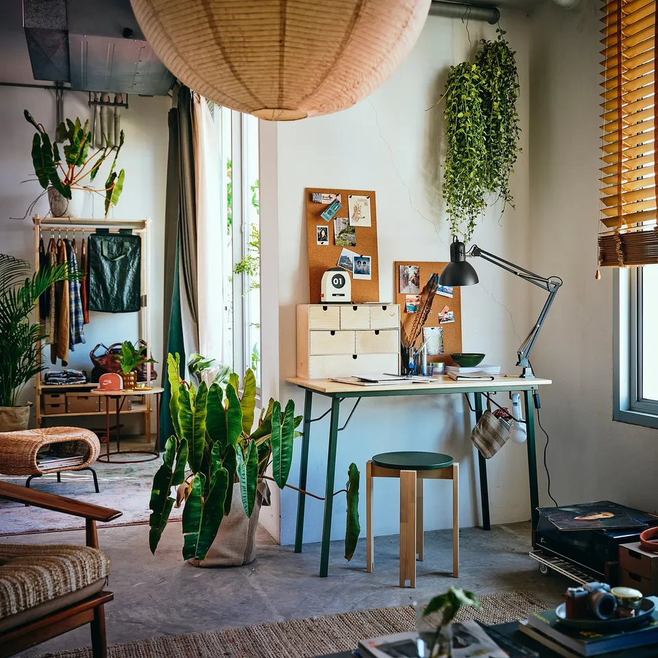 Cosy workspace with KOMPISHÄNG table in centre, surrounded by plants and decor, featuring a large lamp and notice board.