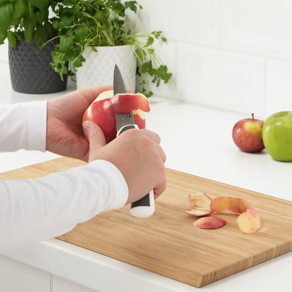 Person peeling apple with paring knife, one of three-knife set, on wooden chopping board with whole apples nearby.