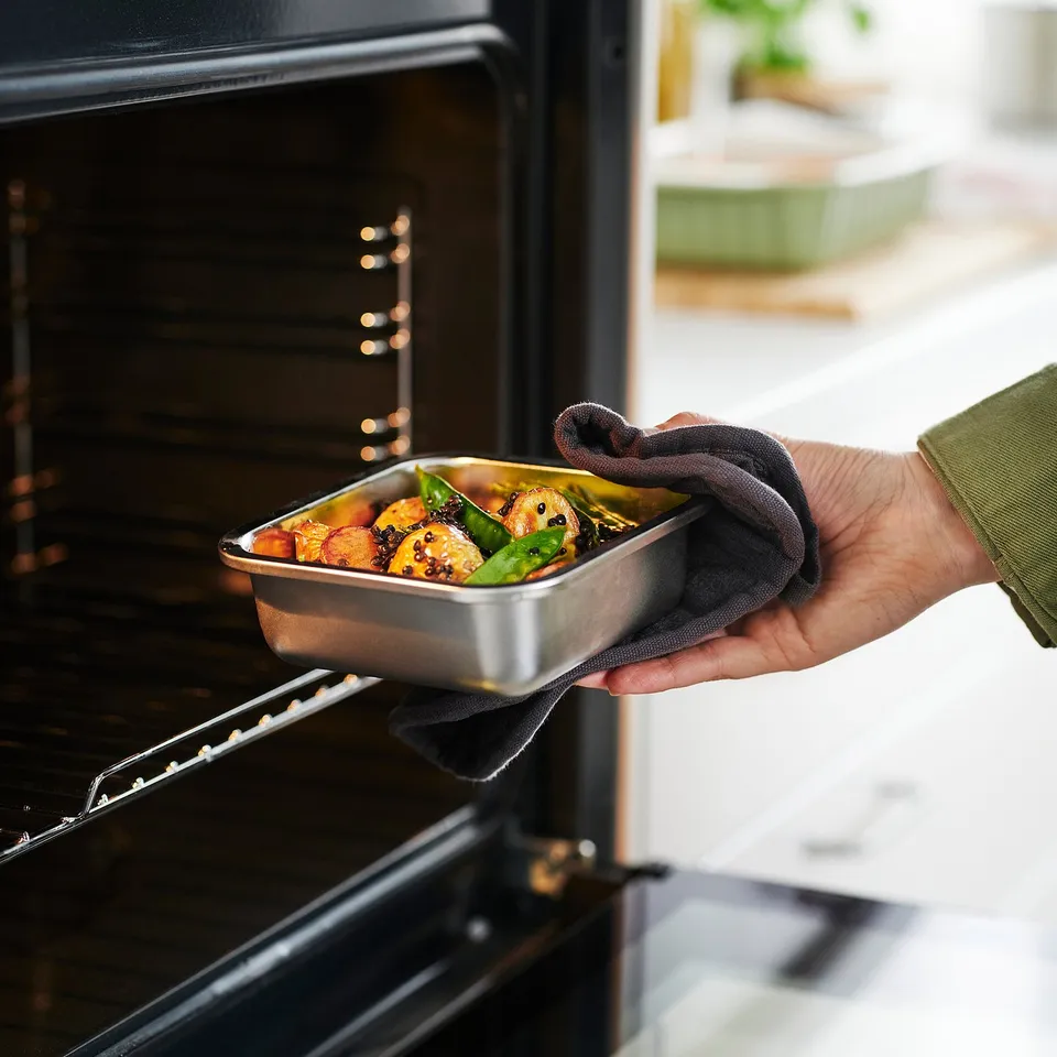 A person places a stainless steel tray of roasted vegetables into an oven, demonstrating the containers oven-safe functionality.