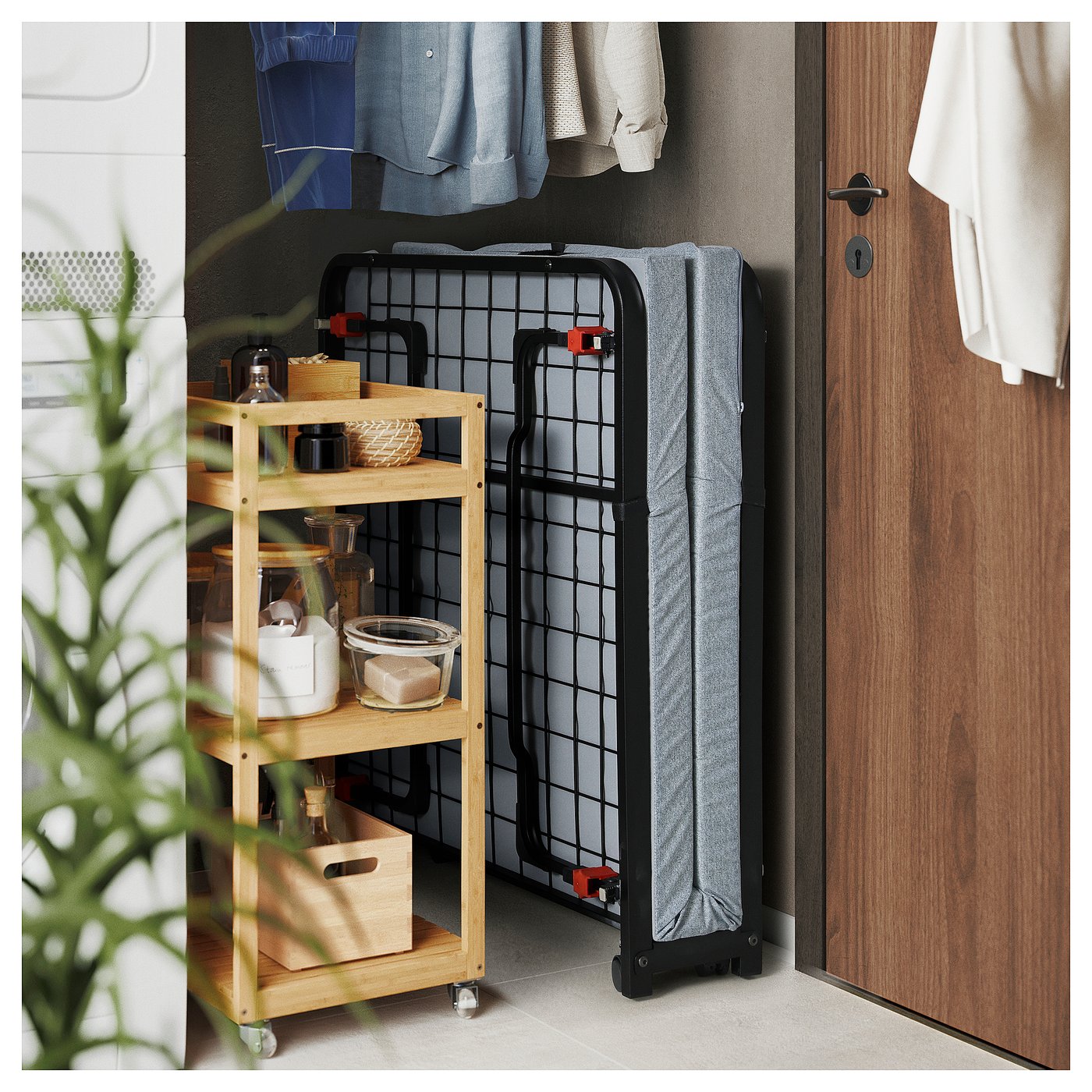 Foldable guest bed, black, leaned against wall near wooden door, next to wooden shelf with kitchen items.