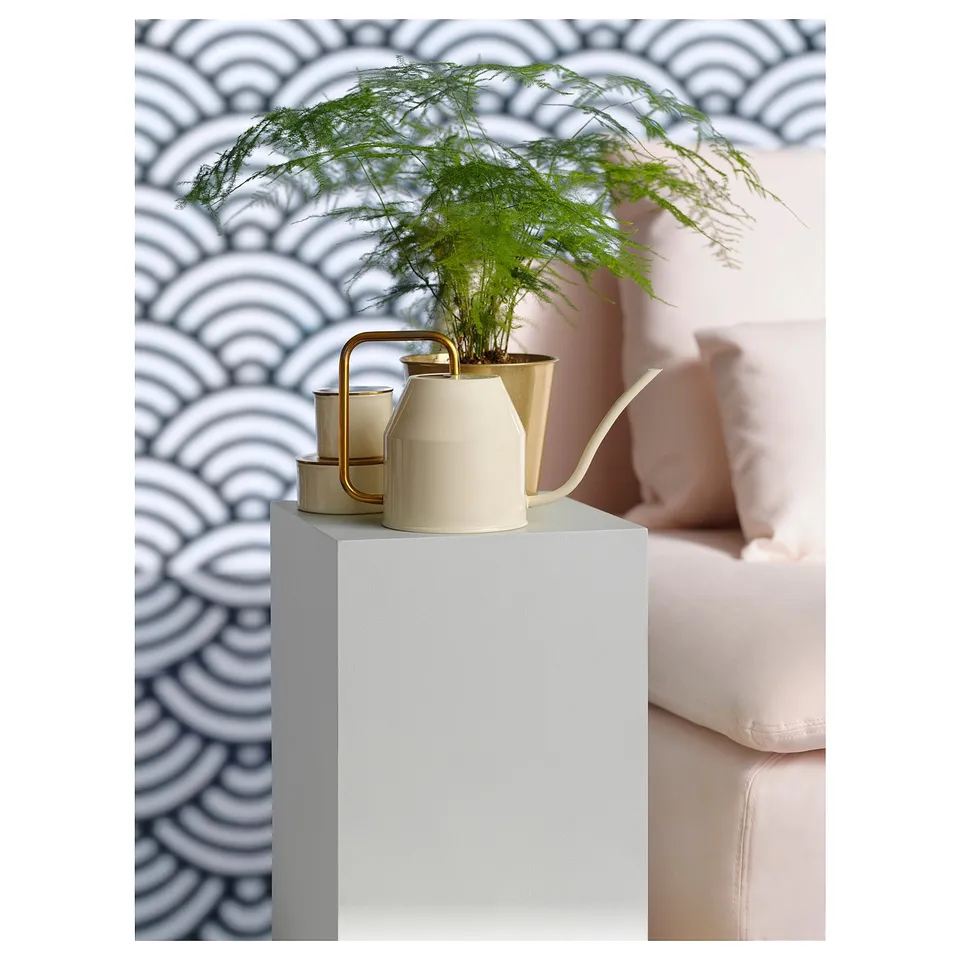 The VATTENKRASSE watering can in beige, made of galvanised steel, sits on a pedestal with a green plant.