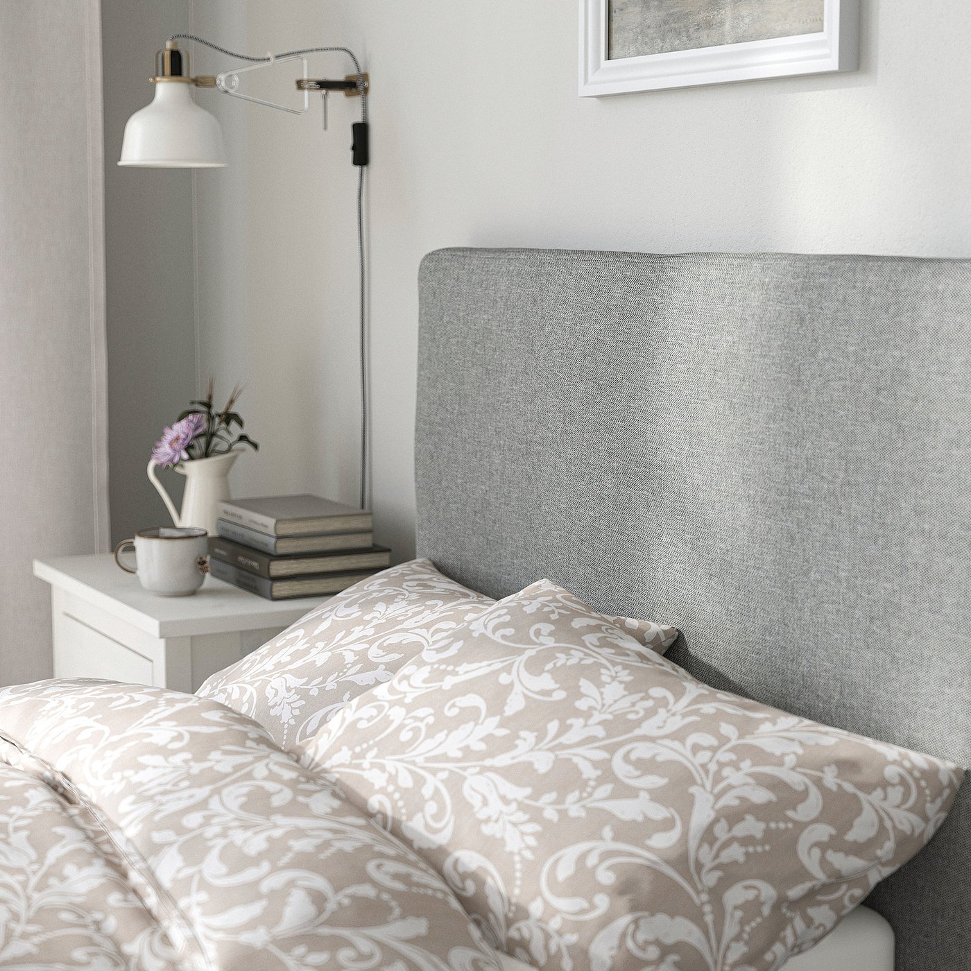 Bedroom scene: grey upholstered headboard, floral bedding, bedside table with lamp, books, and vase.