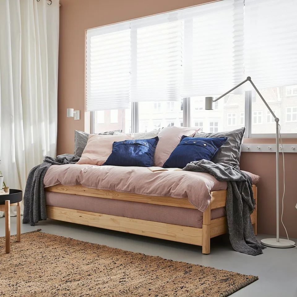 Modern bedroom with large window, wooden daybed, colourful cushions, cosy textiles, and adjustable floor lamp.