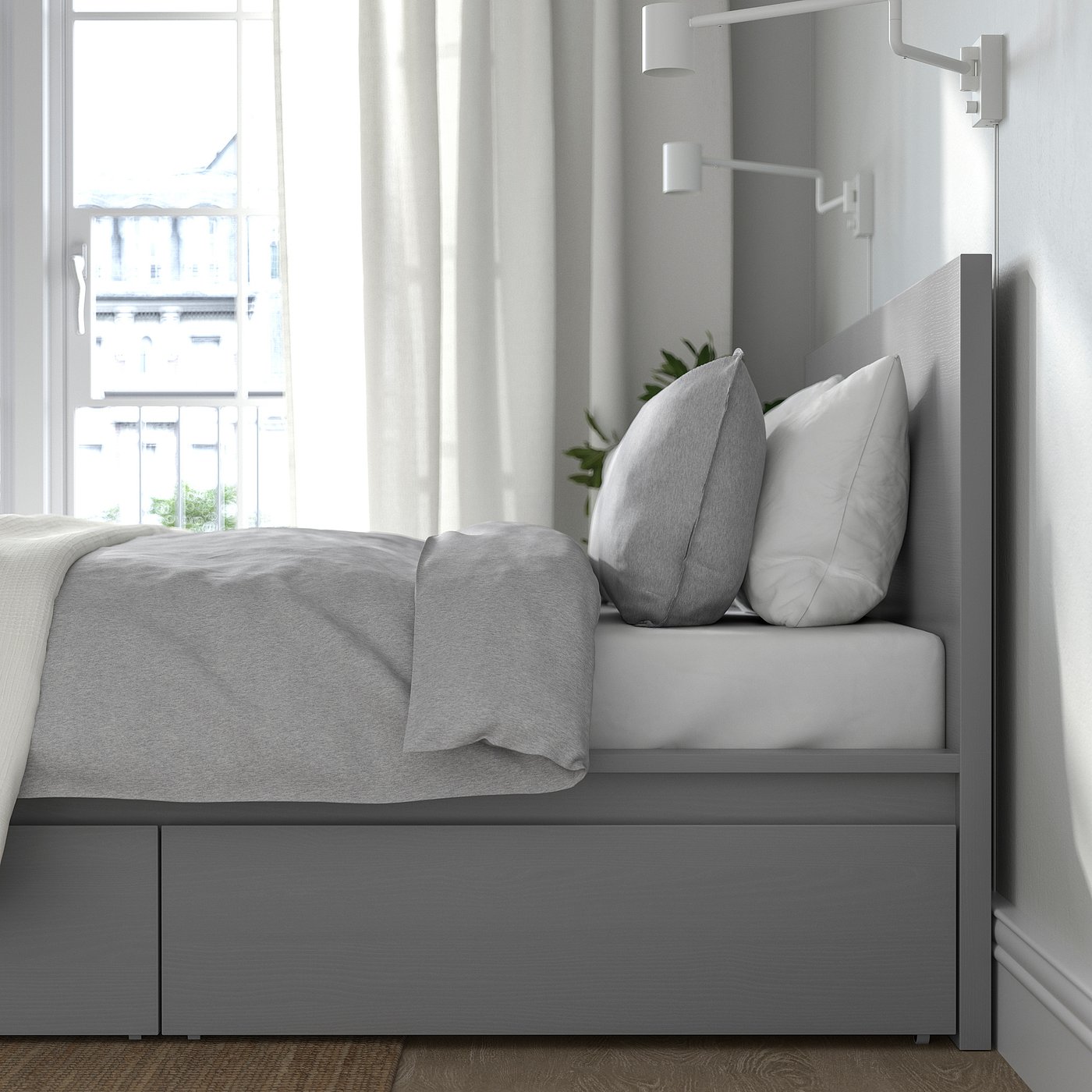 Modern grey bed with drawers and pillows, against a window with white curtains.