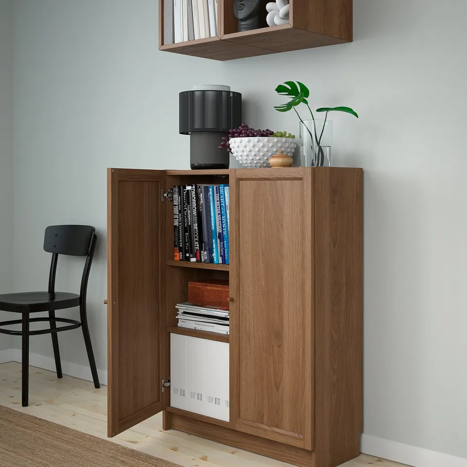 Wooden cabinet with shelves, open doors showing books and magazines, lamp, vase on top, black chair beside.