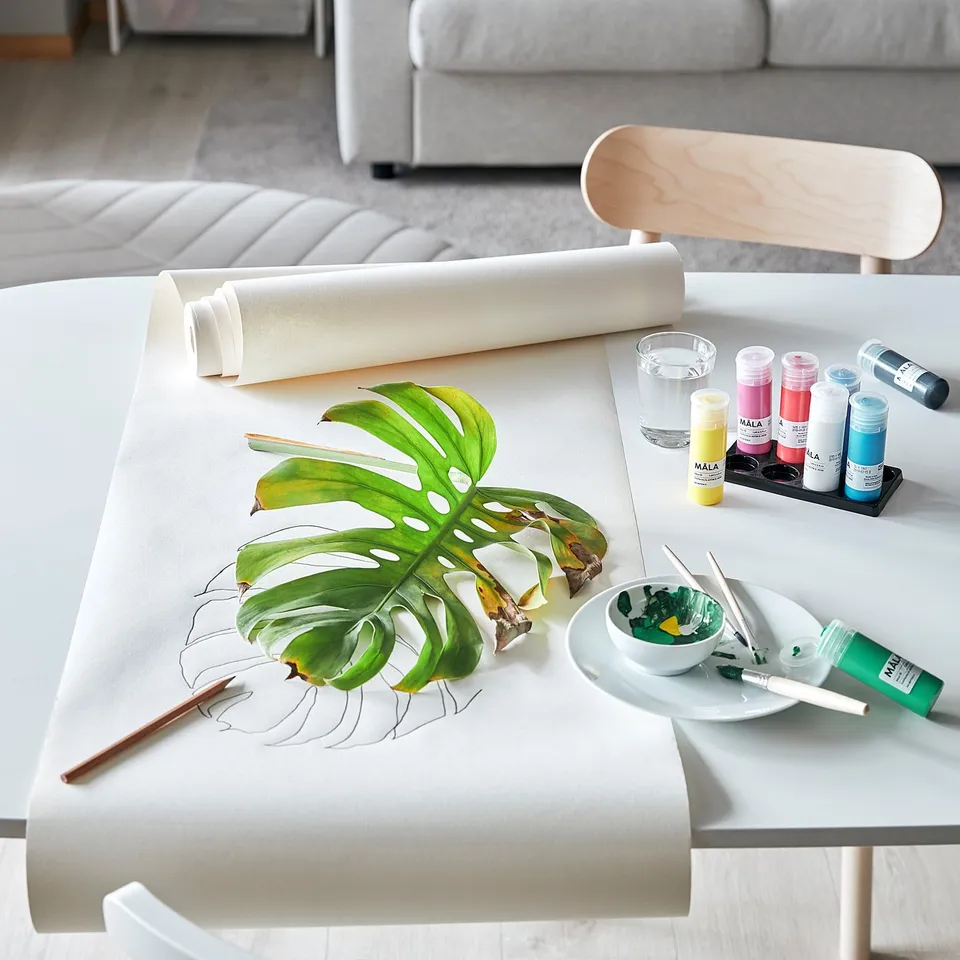 A creative workspace with MÅLA art supplies, featuring a rolling canvas, a monstera leaf, and brushes on a white table with light grey chairs.