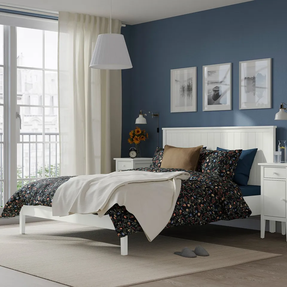 Bedroom with white bed, floral bedding, blue pillows, slippers, window, wall art.