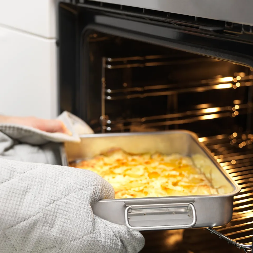 Person removes baked dish from oven using oven mitts. Golden-brown casserole is in a stainless steel baking tray with handles.