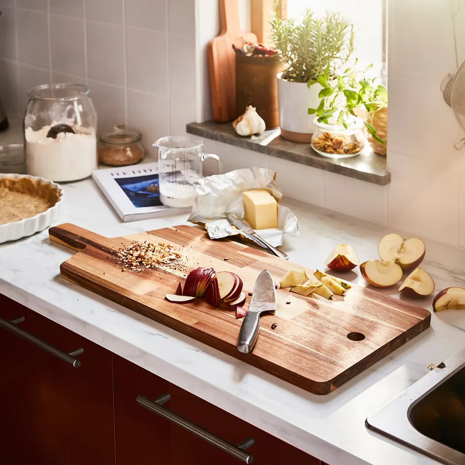 A kitchen counter with a wooden chopping board, knife, sliced apples, butter, measuring cup, and potted plants.