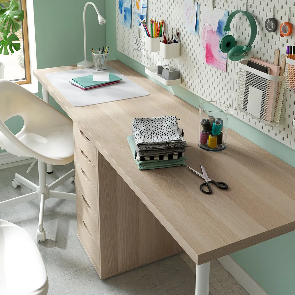 Wooden desk with drawers, notebooks, scissors, threads, wall-mounted pegboard with items, and a white chair.
