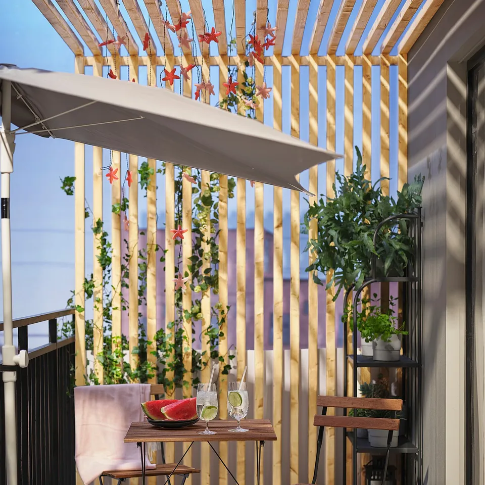 Balcony with TÄRNÖ table & chairs, holding wine & watermelon, surrounded by wood & plants, under awning.