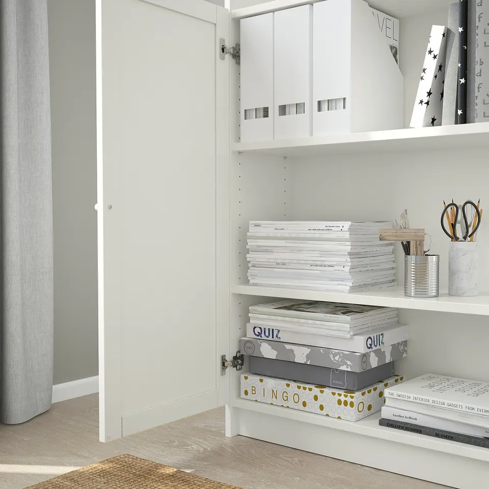 Open white cabinet with organised shelves: boxes, books, stationery, and decorative items arranged neatly within.