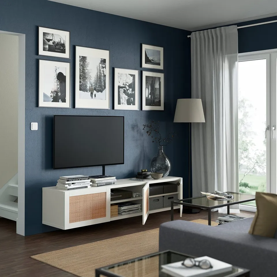 Modern living room: tv on white BESTÅ stand, black framed photos on dark wall, glass coffee table, grey curtains, lamp, and grey sofa.