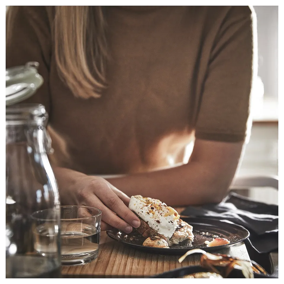 Person holding food on brown plate, glass, and bottle on wooden table. Soft lighting, cosy ambiance.