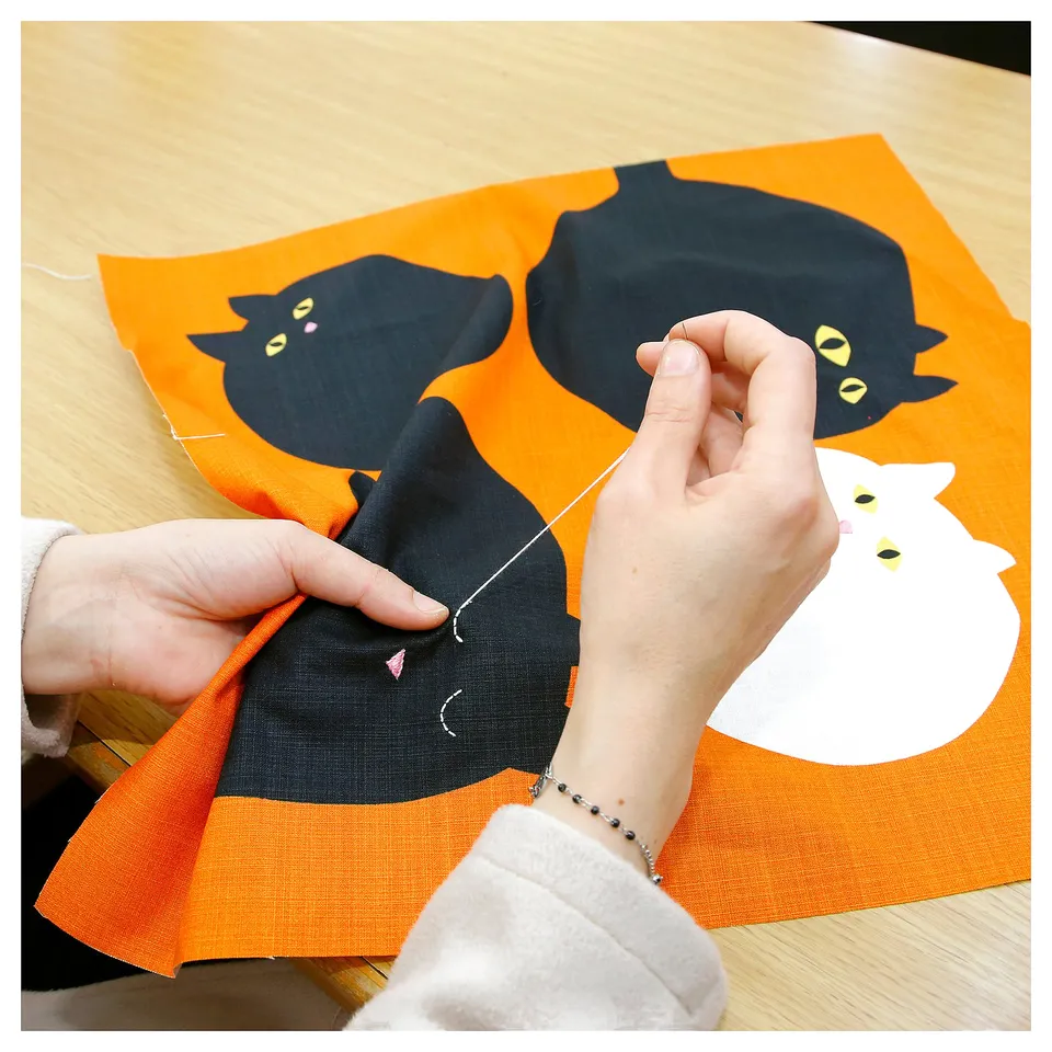 Person stitching cat-themed orange cushion cover with black cats.