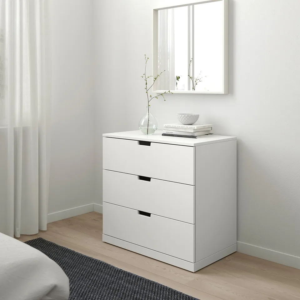 Modern white NORDLI chest of 3 drawers, tall & narrow, next to bed, mirror above, minimalist decor.