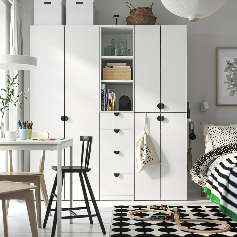 Modern white storage unit with shelves and drawers dominates room, towering over play table and bed.
