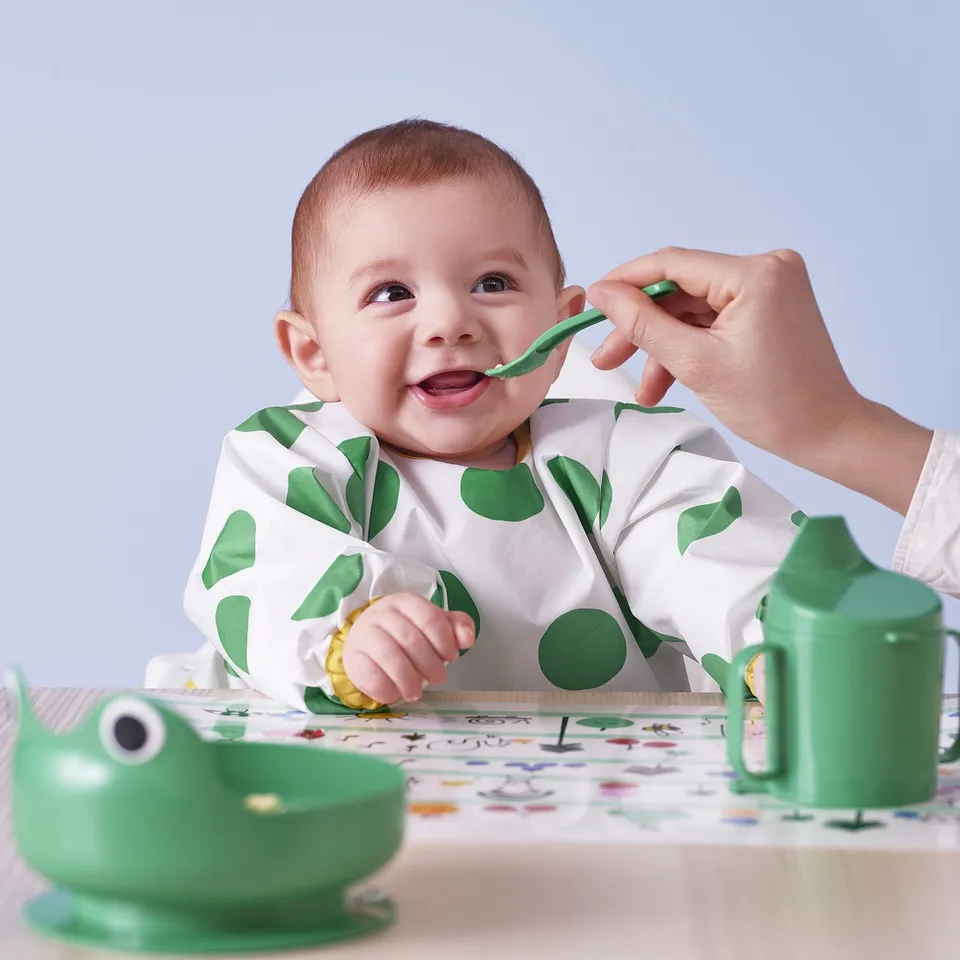 Baby wearing a soft bib with long sleeves, being fed with a green spoon. The bib has a front pocket to catch food and has adjustable.