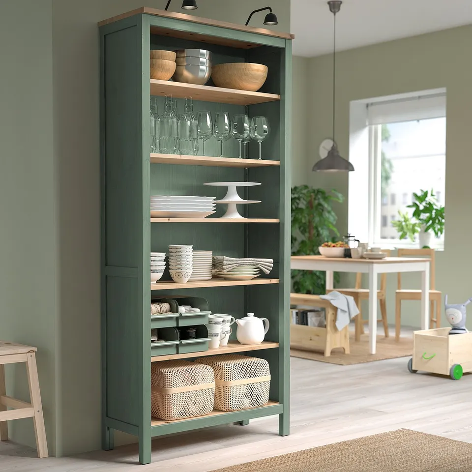 Green HEMNES bookcase with wooden shelves, stacked dishes, and wine glasses, taller than nearby light wooden chair.