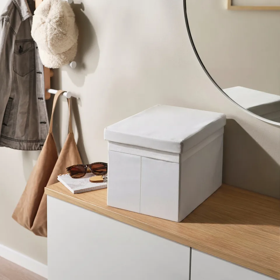 A white fabric box with lid on a wooden shelf, holding sunglasses and a book. The box has integrated handles for easy access.