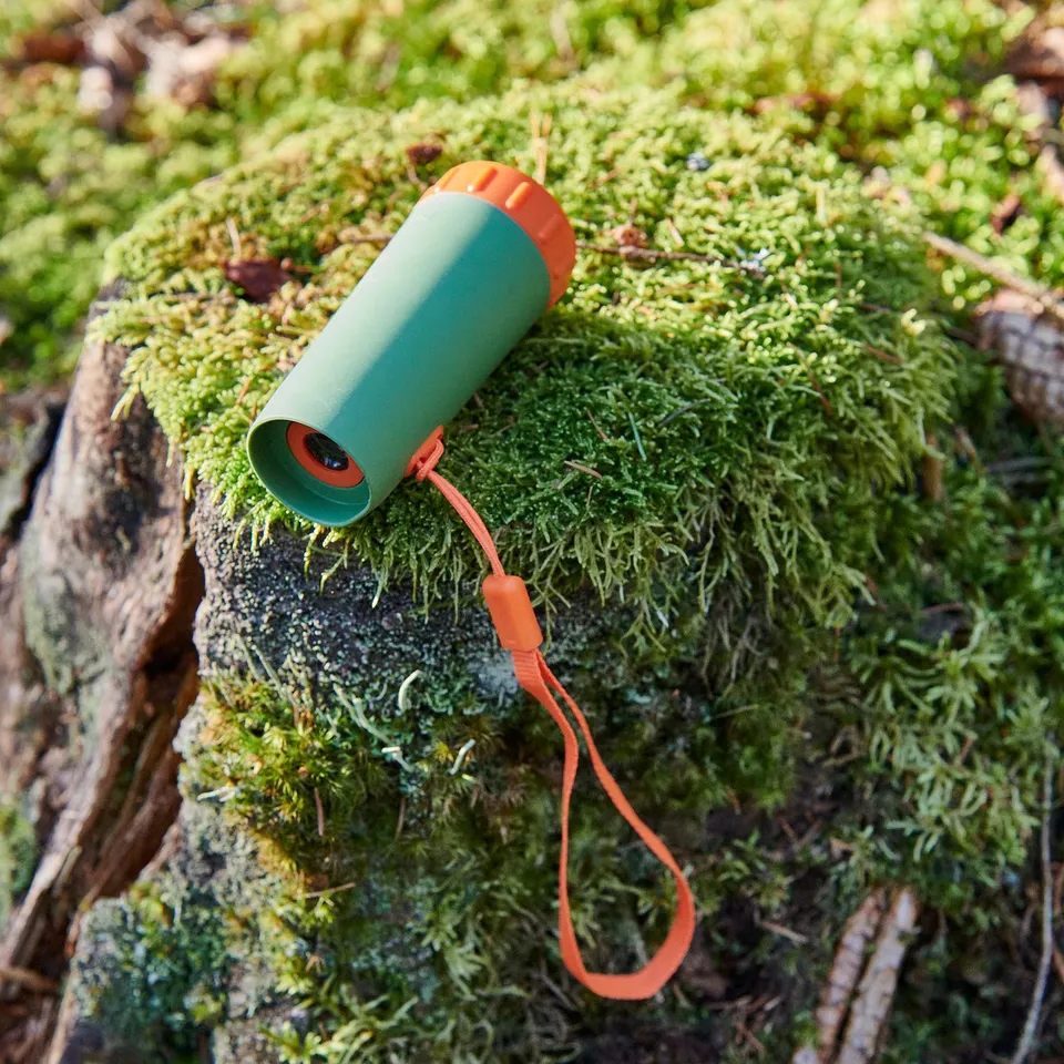 Green and orange monocular rests on mossy log. Bright orange strap attached.