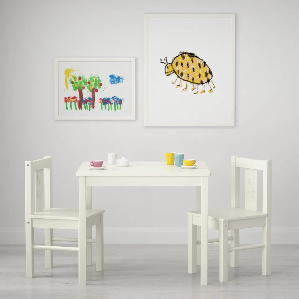 Small white kids table with two chairs, set for tea, under colourful art prints.