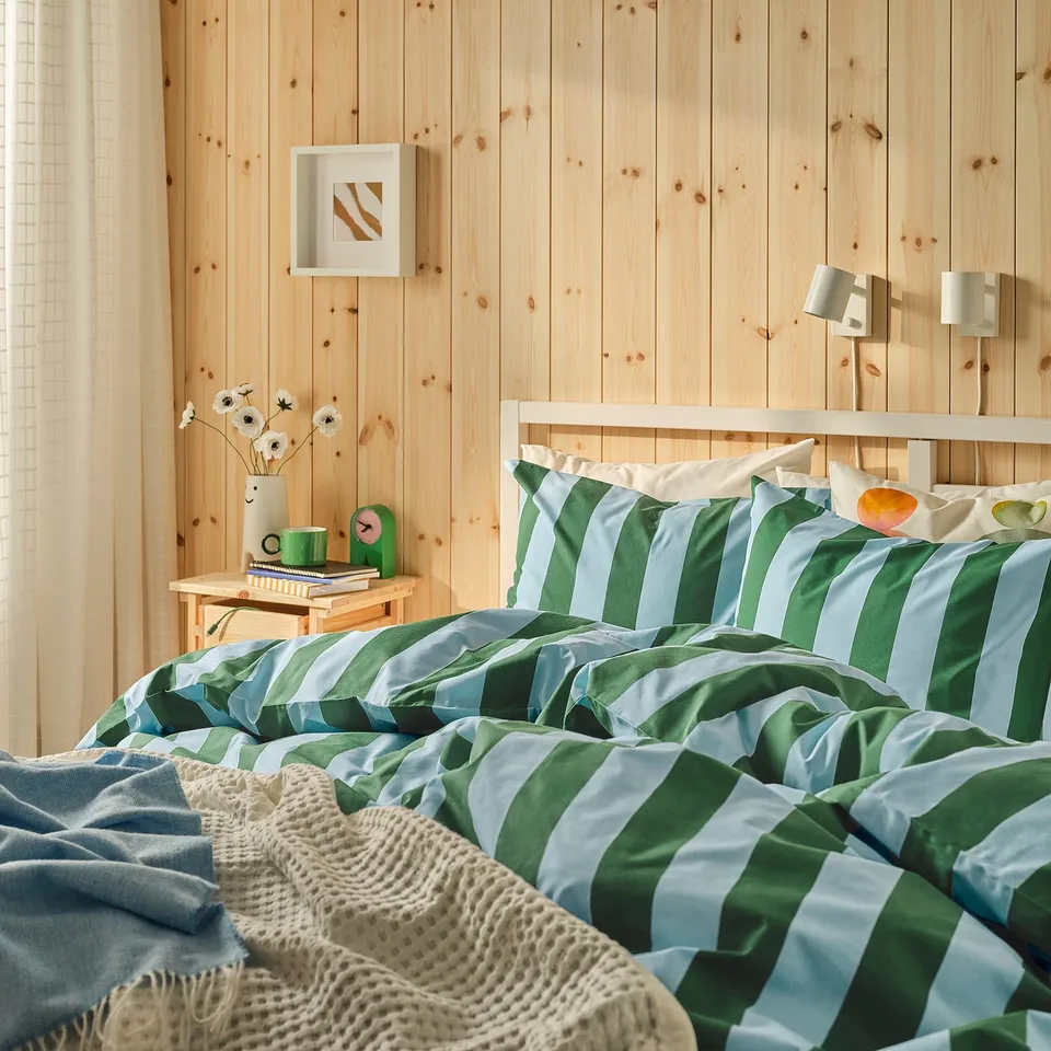 Modern bedroom with SLÅNHÖSTMAL duvet set in green/blue on neatly made bed against wooden wall; small bedside table with vase, clock, books.