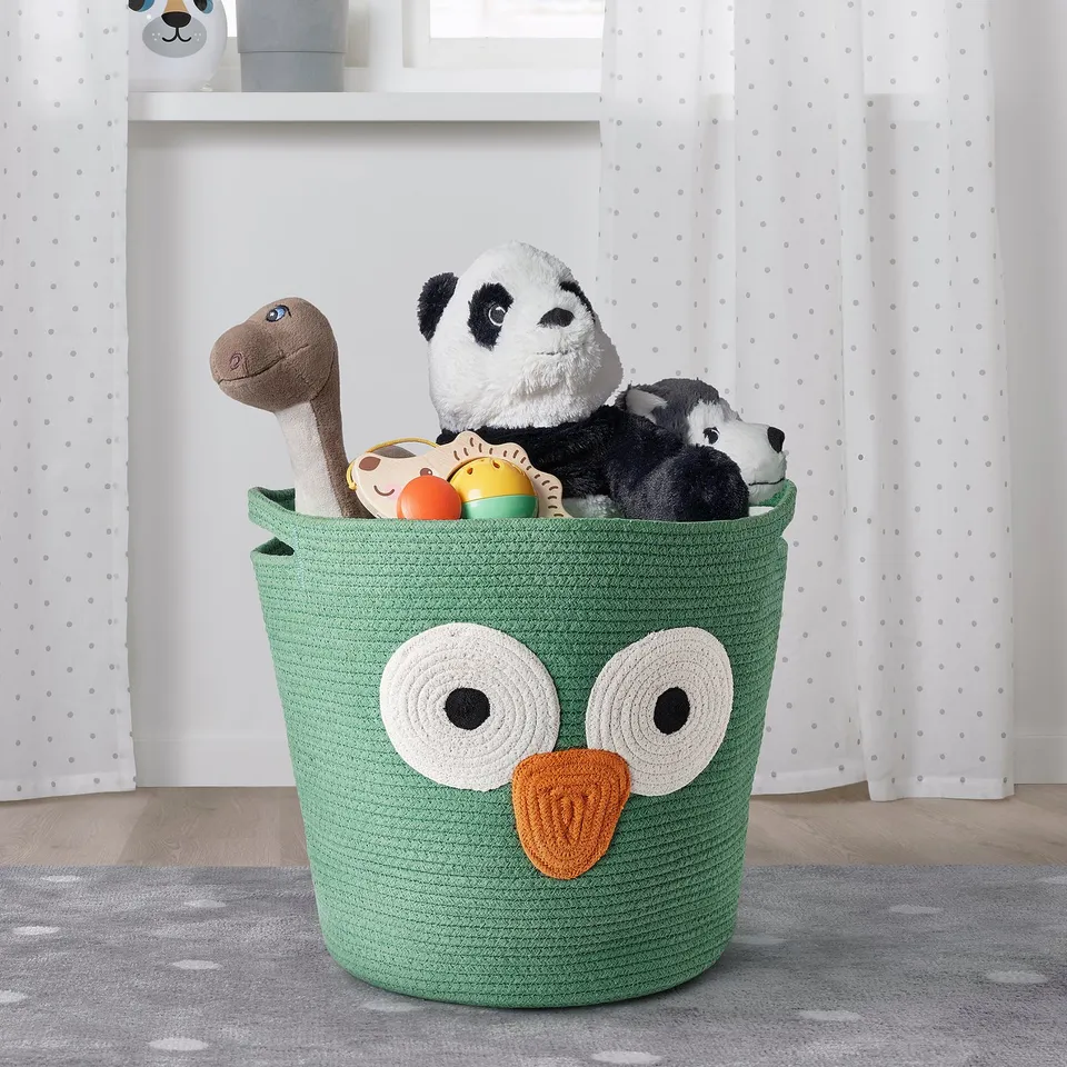 Large green UPPSTÅ storage basket with owl design, filled with toys. Soft cotton material, sits on a grey rug in a bright room.