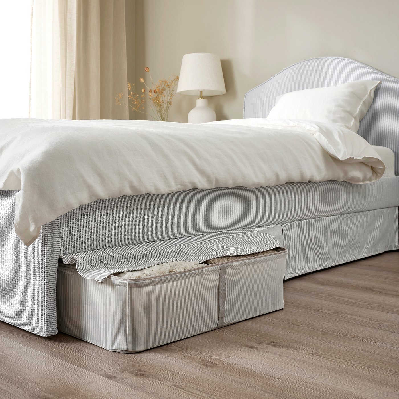 Neat bed with striped frame, white blankets, and open storage at the foot. Beside it, a lamp and dried flowers.