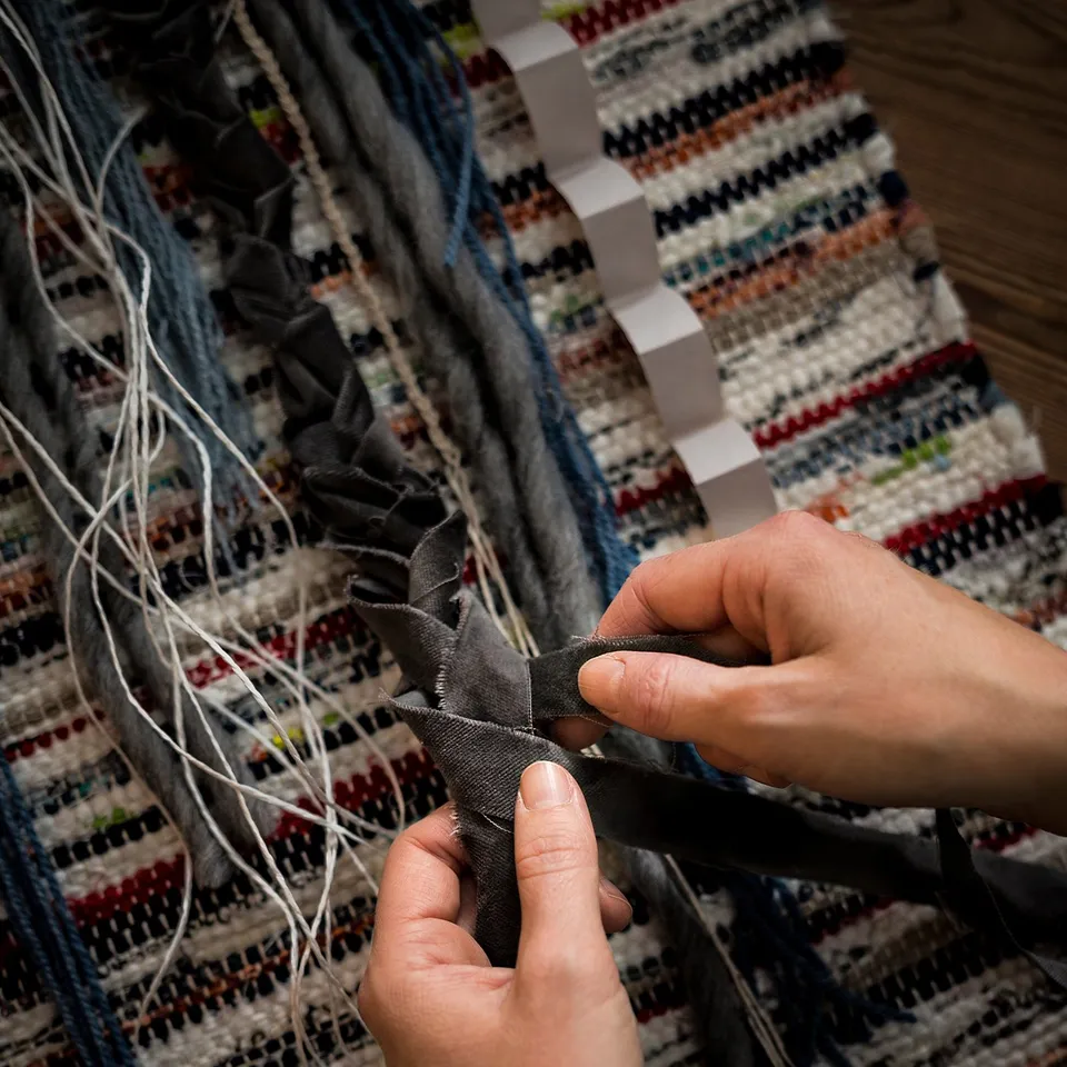 A person is crafting a TÅNUM rug from black cotton strips. The rug intricate patterns on a multi-colored woven base.