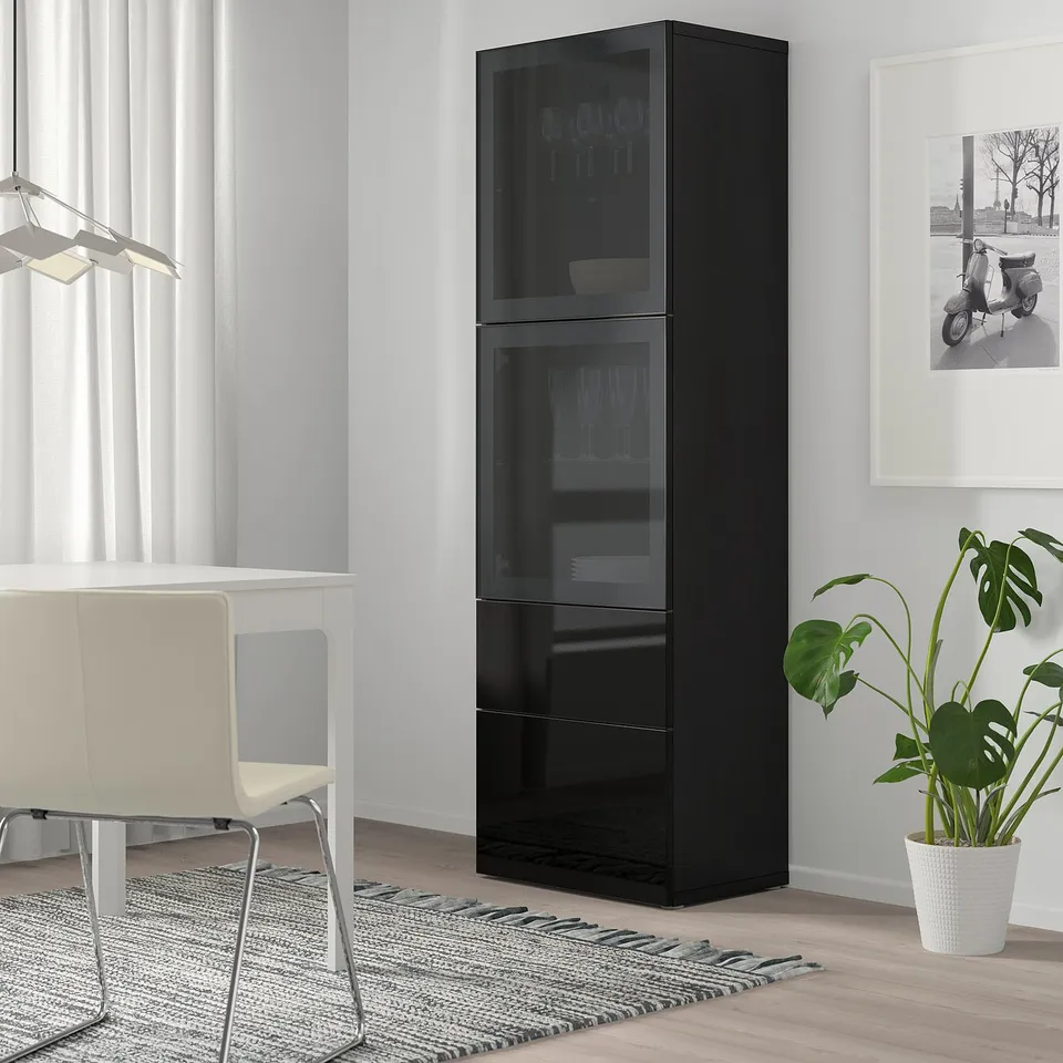 Black BESTÅ storage unit with glass doors next to a white chair and desk in a bright room.