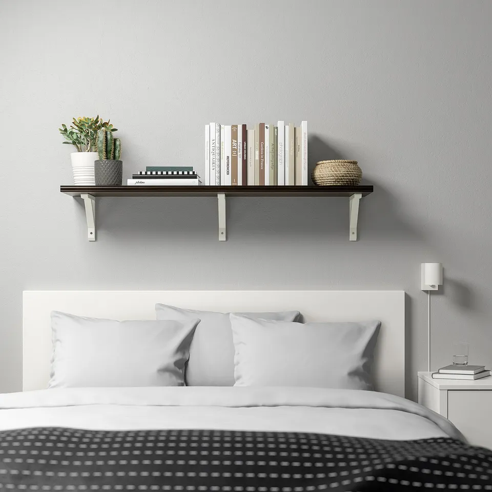 BERGSHULT shelf above bed with books, plant, basket. Dark brown, smooth edges. White headboard, grey pillows, small lamp.