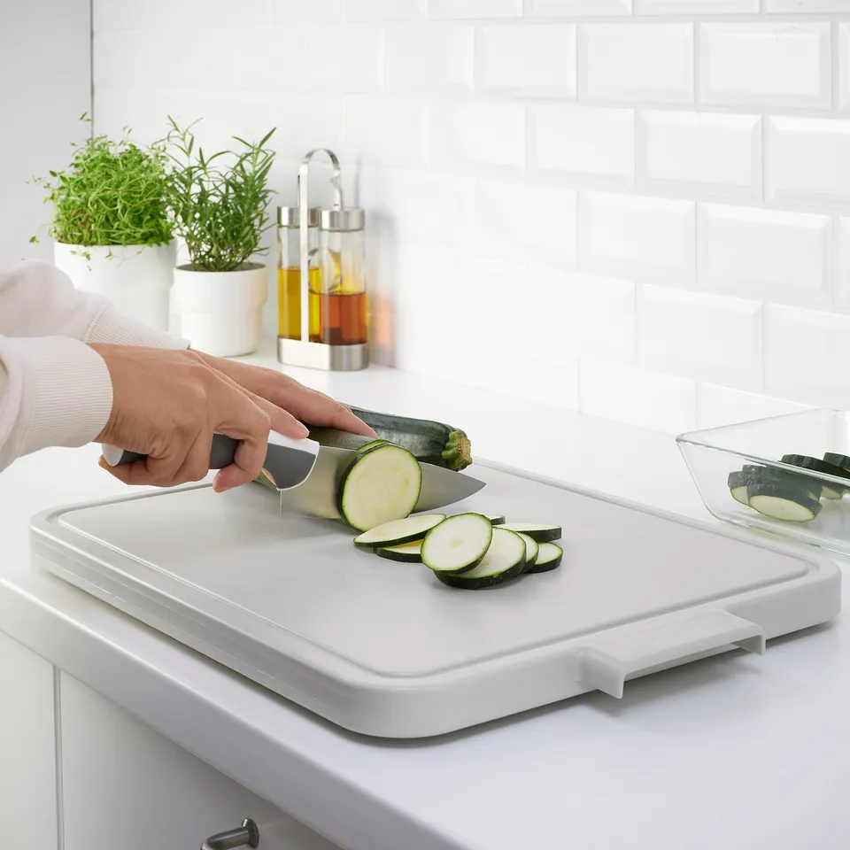 Person slicing courgette on white LILLHAVET chopping board. Cut pieces in plastic container beside.