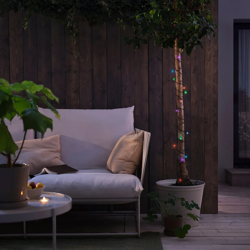 Outdoor seating area with GETKÅL string lights around a tree, white couch, coffee table, and plant for ambiance.