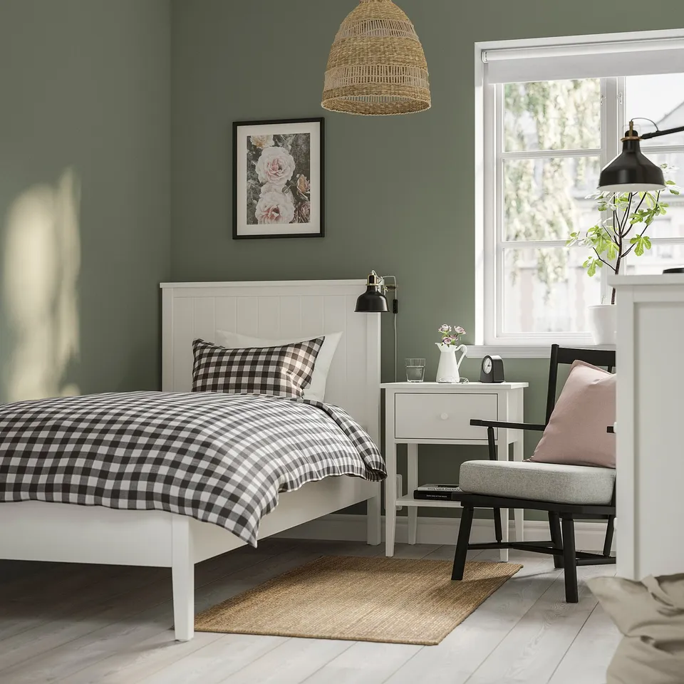 Cosy bedroom: white bed, black/white spread, small table, clock, light, grey chair, rug, green walls, window, lamp.