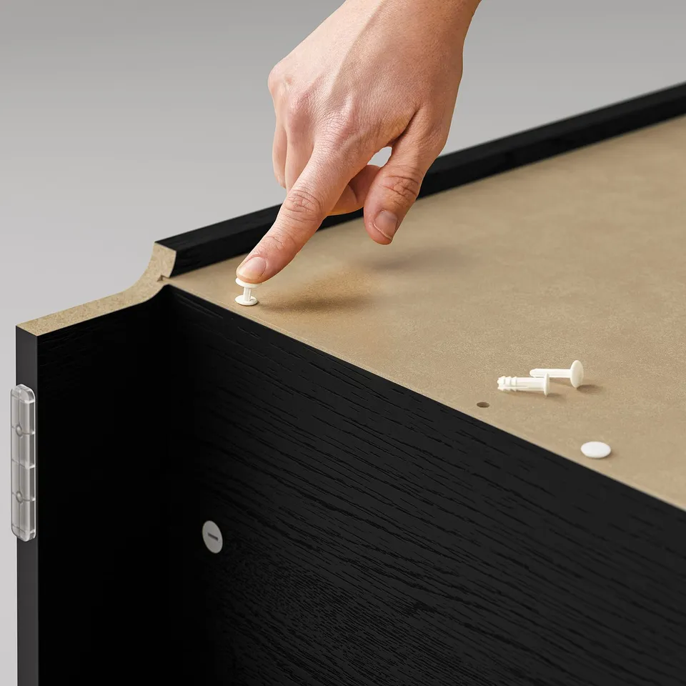 A hand placing a white dowel into a black BILLY bookcase with beige shelves.